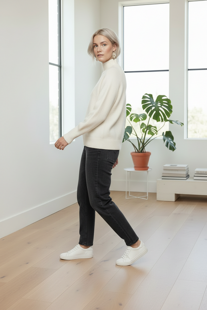 A woman stands in a bright, modern room wearing a cream mock-neck knit sweater, dark charcoal gray jeans, and white sneakers.