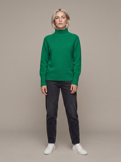 A woman modeling a bright green ribbed turtleneck sweater, paired with dark charcoal gray jeans and white sneakers, standing against a solid warm gray studio background.