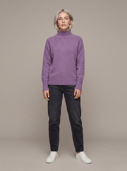 A woman stands against a plain beige studio background wearing a heathered purple mock-neck knit sweater, dark charcoal jeans, and white sneakers.