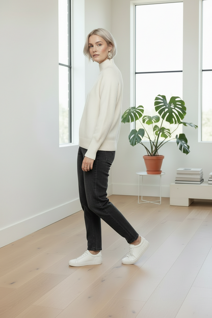 A woman wearing a cream mock-neck knit sweater, black slim-fit jeans, and white sneakers walks across a light wood floor in a bright, modern room with large windows and a potted plant.