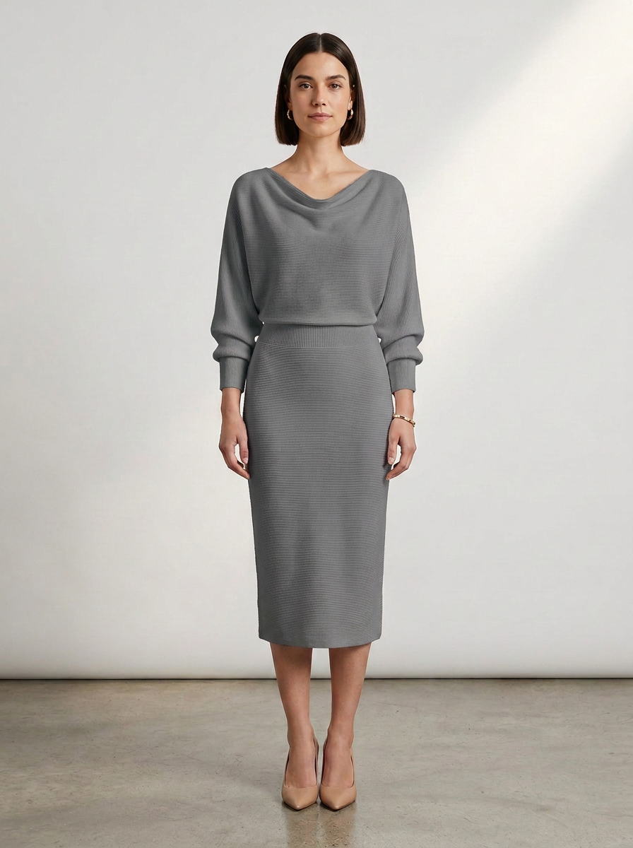 A woman stands in a studio wearing a fitted gray midi dress with long draped sleeves and nude heels against a light gray wall and concrete floor.