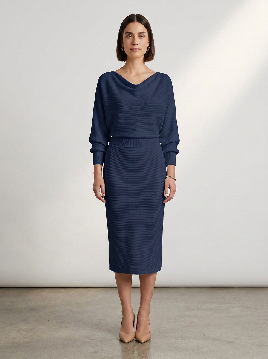 A woman stands in a studio wearing a navy blue long-sleeve midi dress with a draped neckline and a fitted skirt, paired with nude heels against a light gray background.