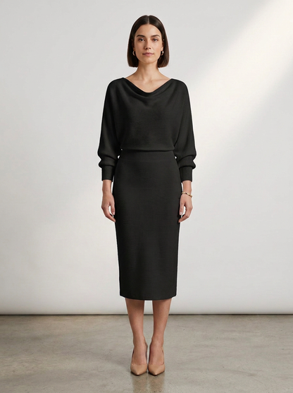 A woman stands in a studio wearing a black long-sleeve midi dress with a draped neckline and a fitted skirt, paired with nude heels against a light gray wall.