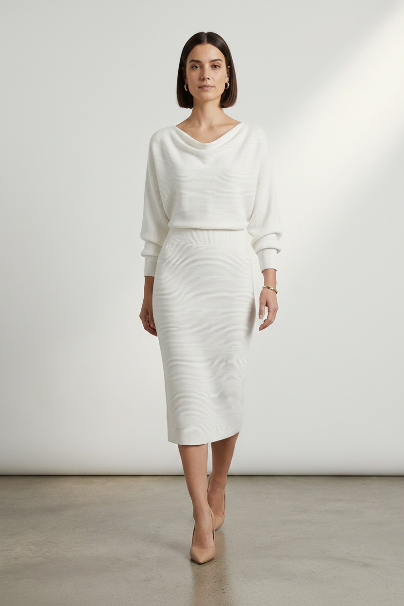A woman stands in a studio wearing a cream-colored knit dress with a draped neckline, long sleeves, and a knee-length skirt. She is wearing nude heels against a clean, light gray background.
