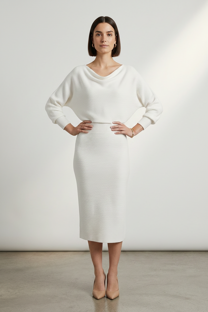 A woman stands in a studio wearing a cream-colored knit dress with a draped neckline, long sleeves, and a knee-length skirt. She is wearing nude heels against a clean, light gray background.