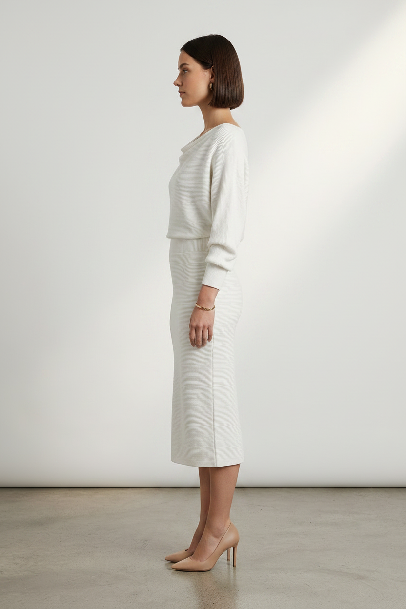 A woman stands in a studio wearing a cream-colored knit dress with a draped neckline, long sleeves, and a knee-length skirt. She is wearing nude heels against a clean, light gray background.