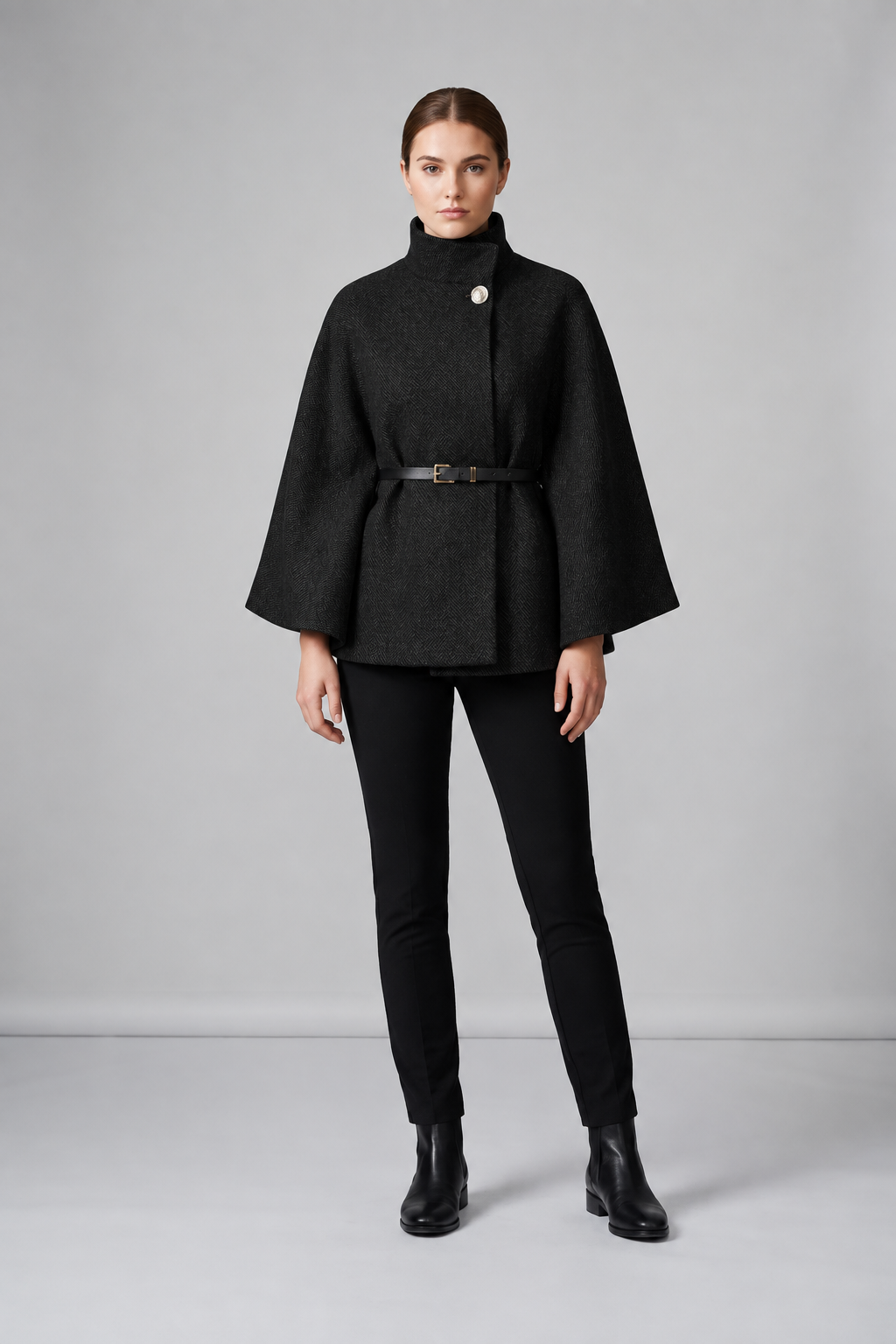 A model stands against a light gray studio background wearing a light gray high-neck cape jacket with a thin black belt, paired with black pants and black ankle boots.