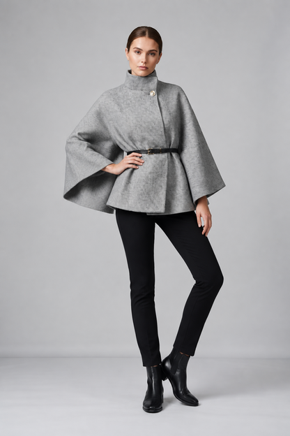 A model stands in a studio wearing a gray high-neck cape-style wrap top with a black belt, black skinny pants, and black ankle boots against a light gray background.