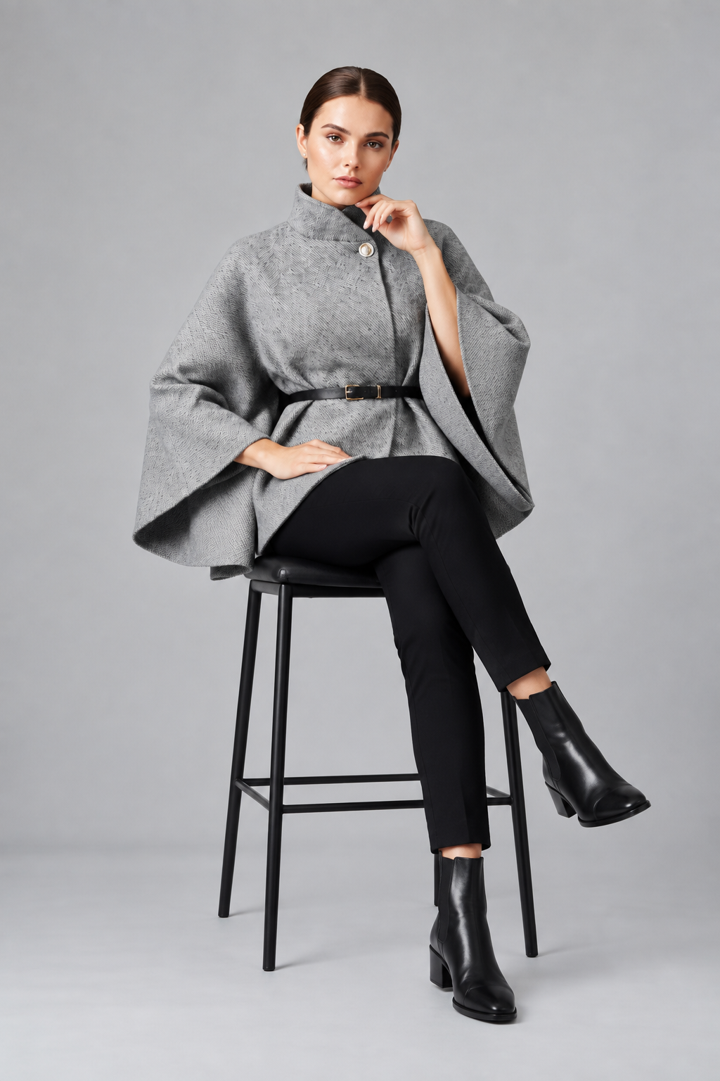 A model stands in a studio wearing a gray high-neck cape-style wrap top with a black belt, black skinny pants, and black ankle boots against a light gray background.