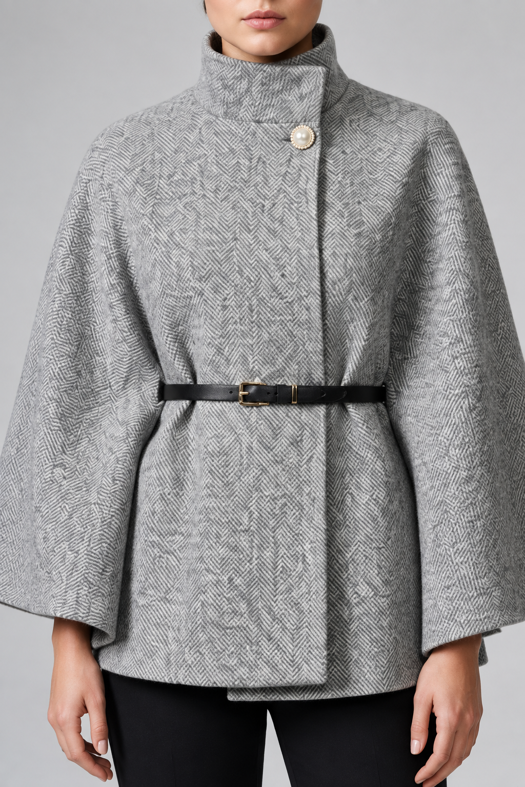 A model wearing a gray and white herringbone patterned cape jacket with a high stand collar, a single decorative button, and a black belt with gold hardware at the waist.