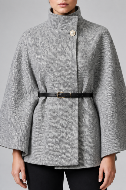 A model wearing a gray and white herringbone patterned cape jacket with a high stand collar, a single decorative button, and a black belt with gold hardware at the waist.