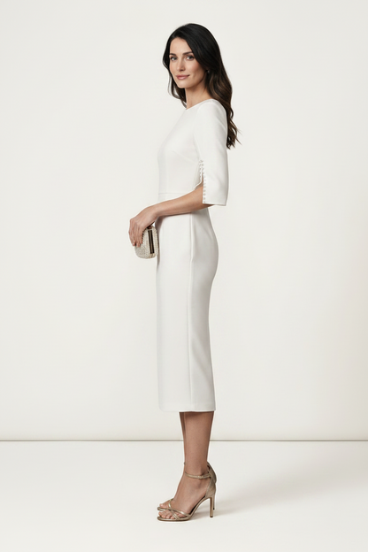 A woman models a white knee-length sheath dress with elbow-length sleeves featuring cutout details on the sides, styled with a small metallic clutch and nude high-heeled sandals against a plain off-white studio background.