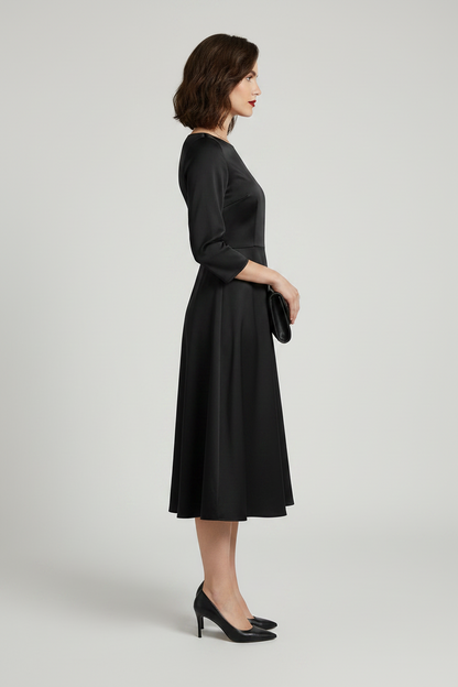 A woman modeling a black three-quarter sleeve fit-and-flare midi dress with a boat neckline, holding a black clutch purse against a light gray studio background.
