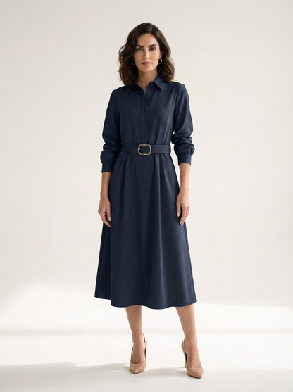 A woman modeling a navy blue midi shirt dress with a matching belt and gold-tone buckle, standing against a clean off-white studio background.