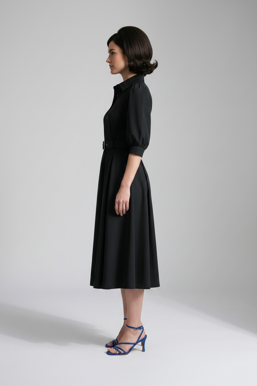 A woman modeling a black midi shirt dress with puff sleeves, a pointed collar, and a matching black belt with a silver buckle, paired with blue strappy heels against a light gray studio background.