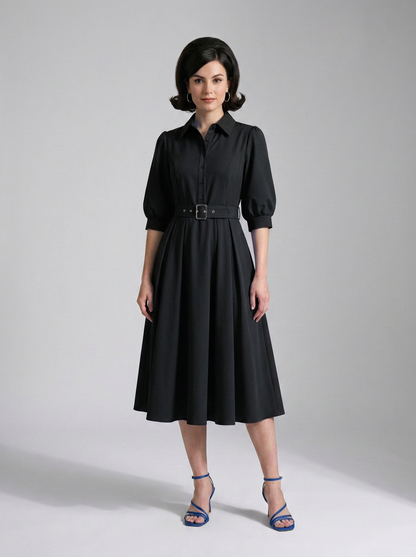 A woman modeling a black midi shirt dress with puff sleeves, a pointed collar, and a matching black belt with a silver buckle, paired with blue strappy heels against a light gray studio background.