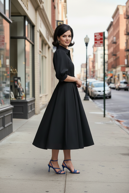 A woman stands on a city sidewalk wearing a black midi dress with puff sleeves, a pointed collar, and a fitted waist, paired with blue strappy high-heel sandals. Buildings and parked cars are visible in the background.