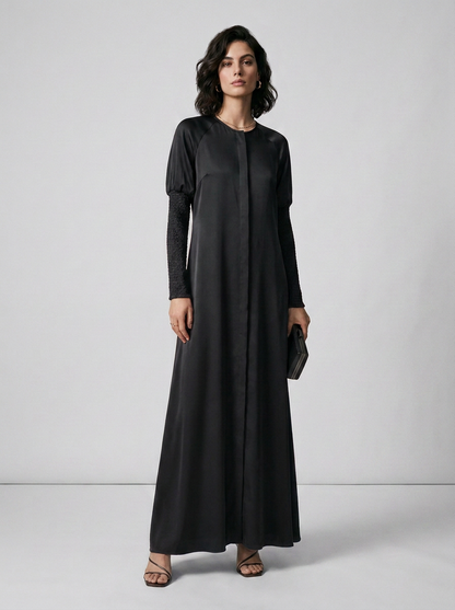 A woman modeling a long black maxi dress with long sleeves and a round neckline, standing against a light gray studio background.