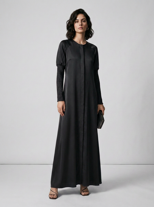 A woman modeling a long black maxi dress with long sleeves and a round neckline, standing against a light gray studio background.
