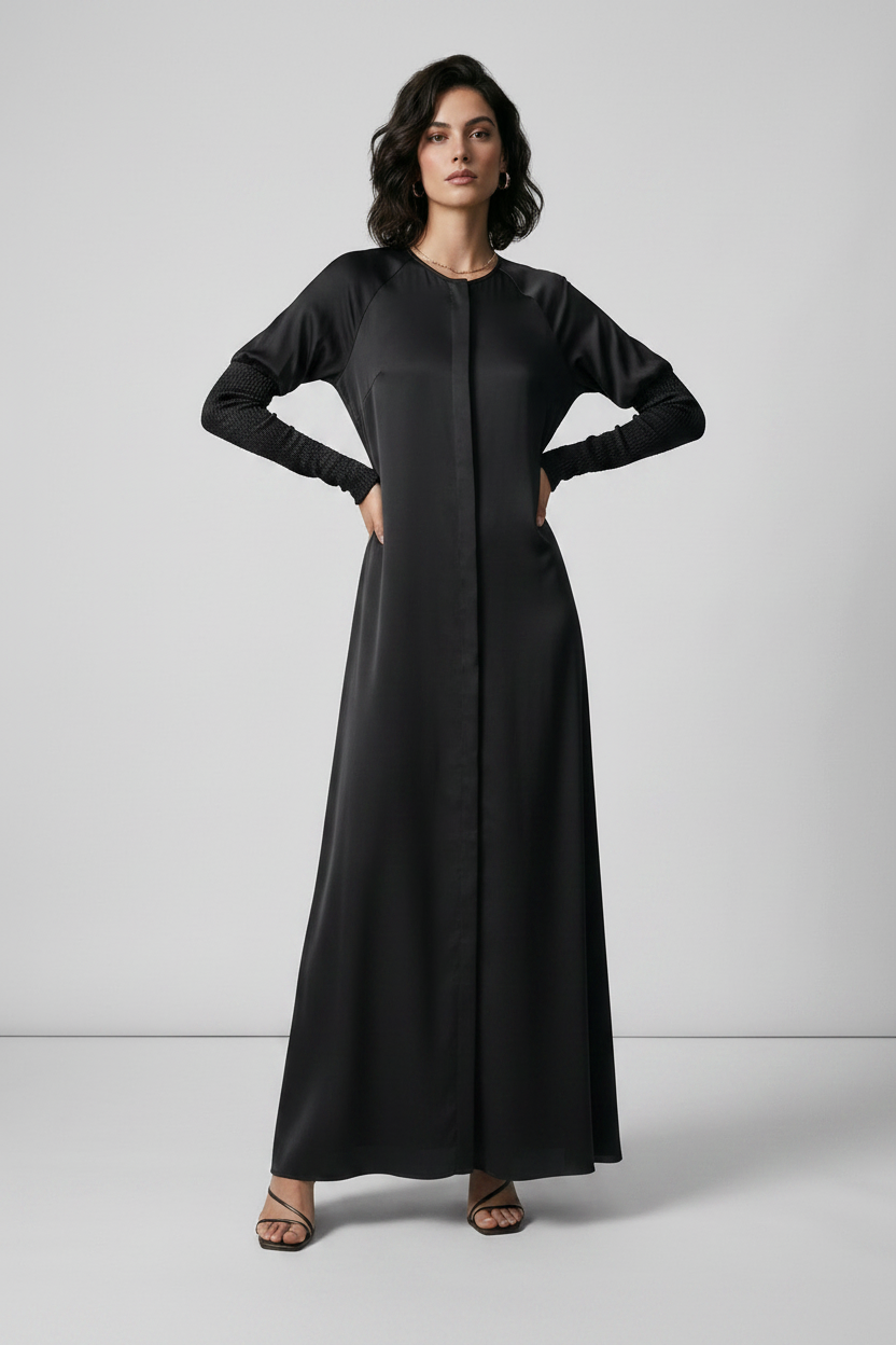 A woman modeling a long black maxi dress with long sleeves and a round neckline, standing against a light gray studio background.