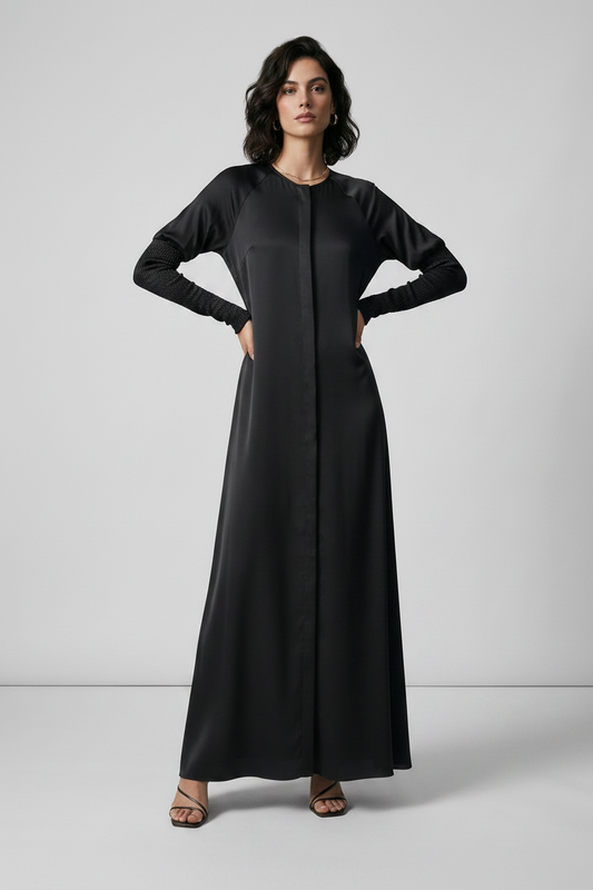 A woman modeling a long black maxi dress with long sleeves and a round neckline, standing against a light gray studio background.