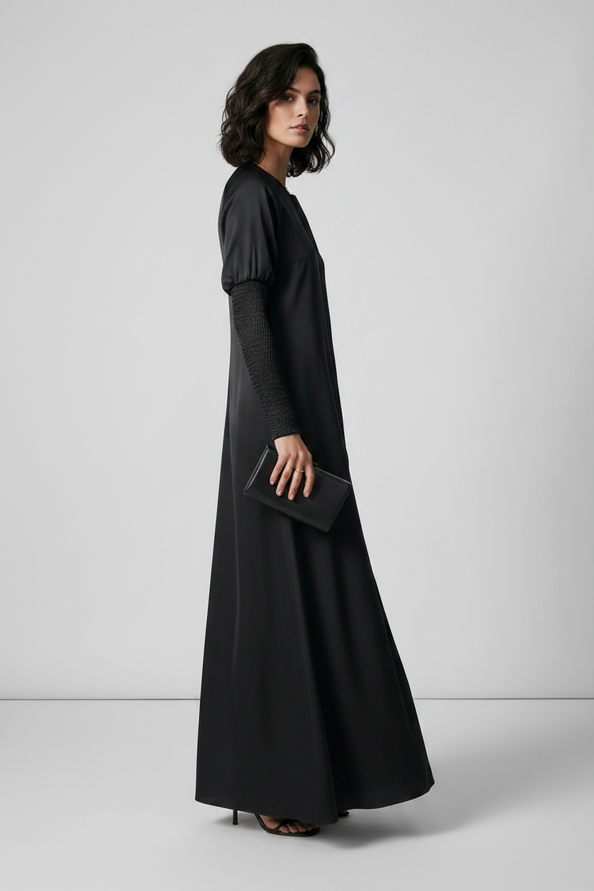 A woman modeling a long black maxi dress with long sleeves and a round neckline, standing against a light gray studio background.