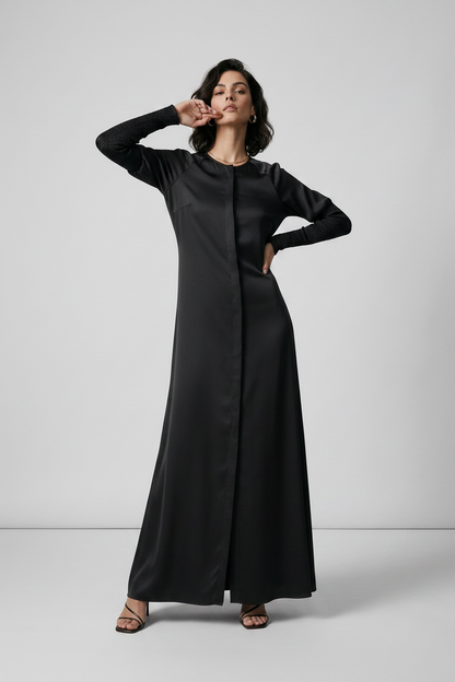A woman modeling a long black maxi dress with long sleeves and a round neckline, standing against a light gray studio background.