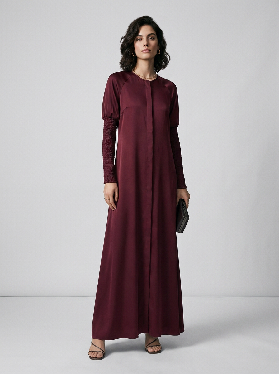 A woman modeling a long-sleeved, floor-length maroon maxi dress with a round neckline and puffed shoulders, standing against a light gray studio background.