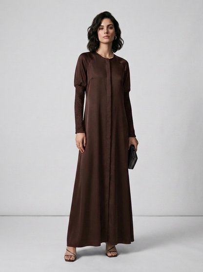 A woman modeling a long-sleeved, floor-length brown dress with a round neckline and a subtle sheen, standing against a light gray studio background.