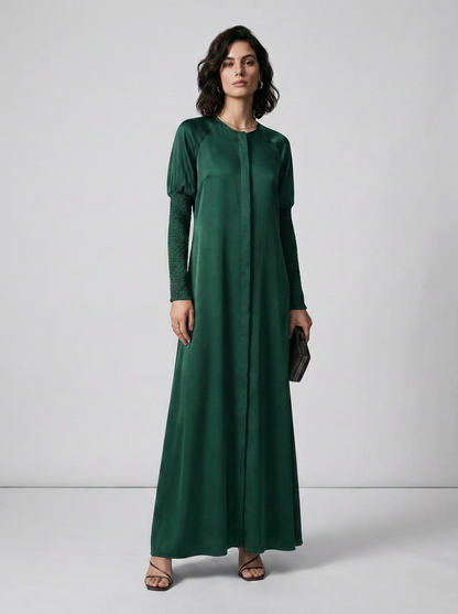A woman modeling a long-sleeved, floor-length emerald green dress with textured sleeves, standing against a light gray studio background.