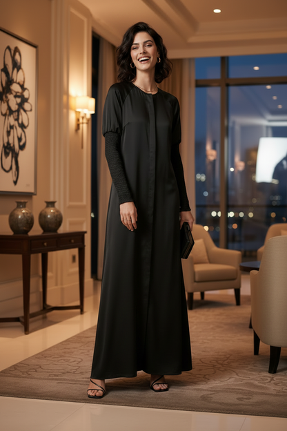 A woman stands in a warmly lit modern interior wearing a long black satin-like dress with long sleeves and a straight, floor-length silhouette. She is holding a small black clutch and wearing flat sandals.