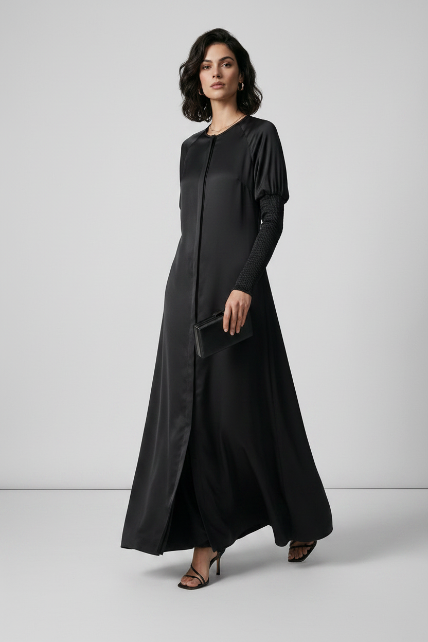 A woman modeling a long black maxi dress with long sleeves and a round neckline, standing against a light gray studio background.