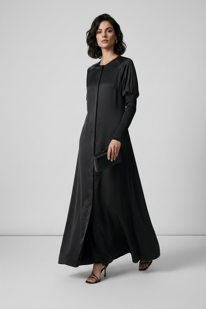 A woman modeling a long black maxi dress with long sleeves and a round neckline, standing against a light gray studio background.