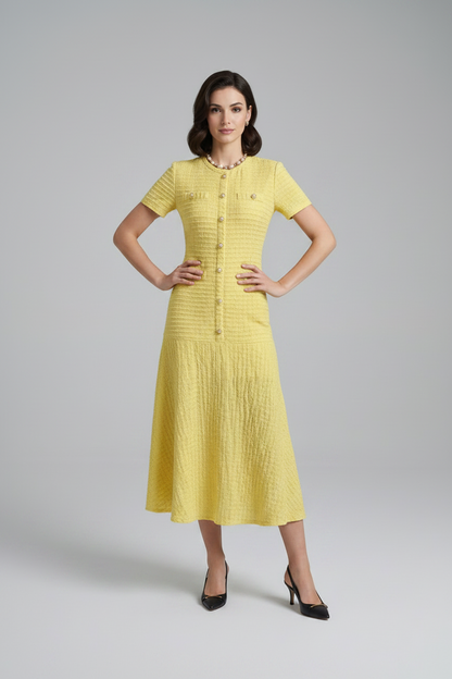 A woman models a textured pale yellow short-sleeve midi dress with a button-front bodice and pleated skirt, styled with a white handbag and black heels against a light gray studio background.
