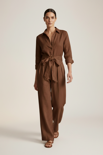 A woman stands against a warm off-white studio background wearing a brown long-sleeve linen jumpsuit with a tie waist and wide legs, paired with brown sandals.