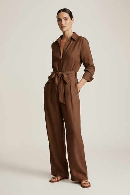 A woman stands against a warm off-white studio background wearing a brown long-sleeve linen jumpsuit with a tie waist and wide legs, paired with brown sandals.