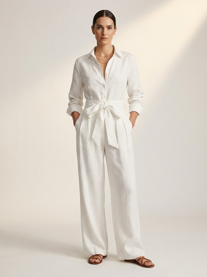 A woman modeling a white long-sleeve linen jumpsuit with a tie waist, standing against a warm off-white studio background.