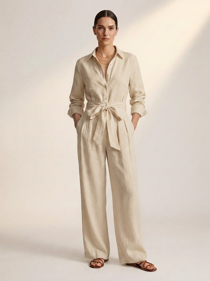 A woman modeling a light beige linen jumpsuit with a tie waist, long sleeves, and wide legs, paired with brown leather sandals against a warm off-white studio background.