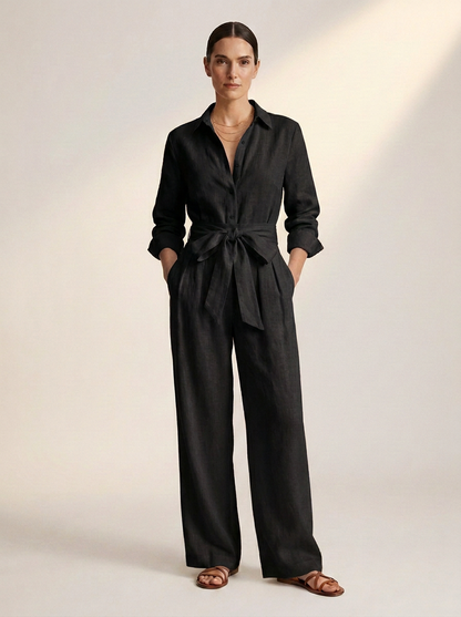 A woman modeling a black long-sleeve linen jumpsuit with a tie waist and wide legs, paired with brown leather slide sandals, against a warm off-white studio background.