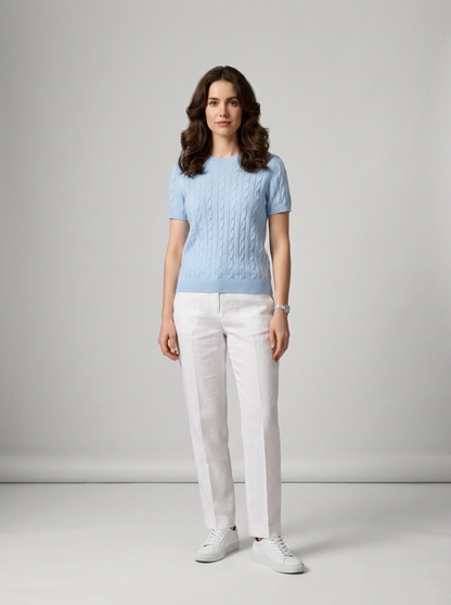 A woman stands in a studio wearing a light blue short-sleeve cable-knit sweater, white trousers, and white sneakers against a light gray background.