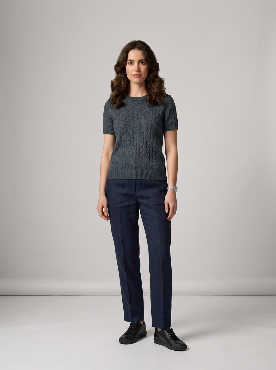 A woman stands in a studio wearing a dark gray short-sleeve cable-knit top, navy blue trousers, and black shoes against a light gray background.
