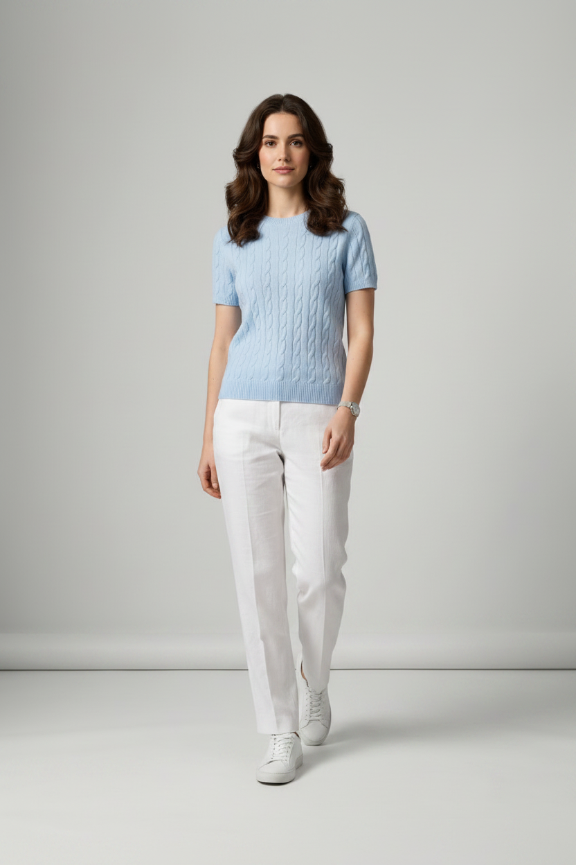 A woman stands in a studio wearing a light blue short-sleeve cable-knit sweater, white trousers, and white sneakers against a light gray background.