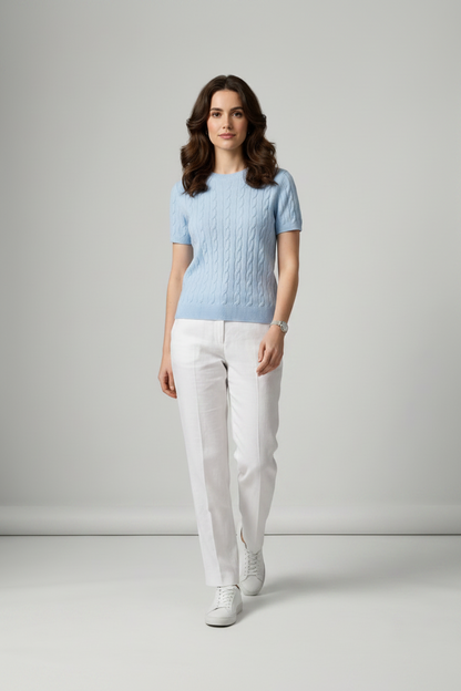 A woman stands in a studio wearing a light blue short-sleeve cable-knit sweater, white trousers, and white sneakers against a light gray background.