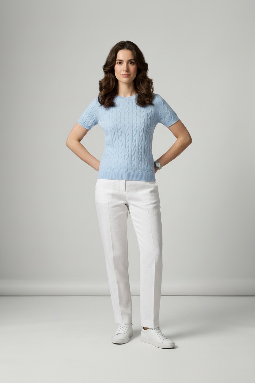 A woman stands in a studio wearing a light blue short-sleeve cable-knit sweater, white trousers, and white sneakers against a light gray background.