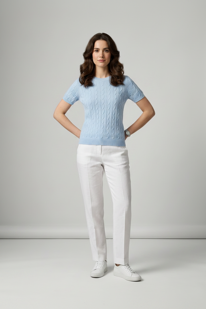 A woman stands in a studio wearing a light blue short-sleeve cable-knit sweater, white trousers, and white sneakers against a light gray background.