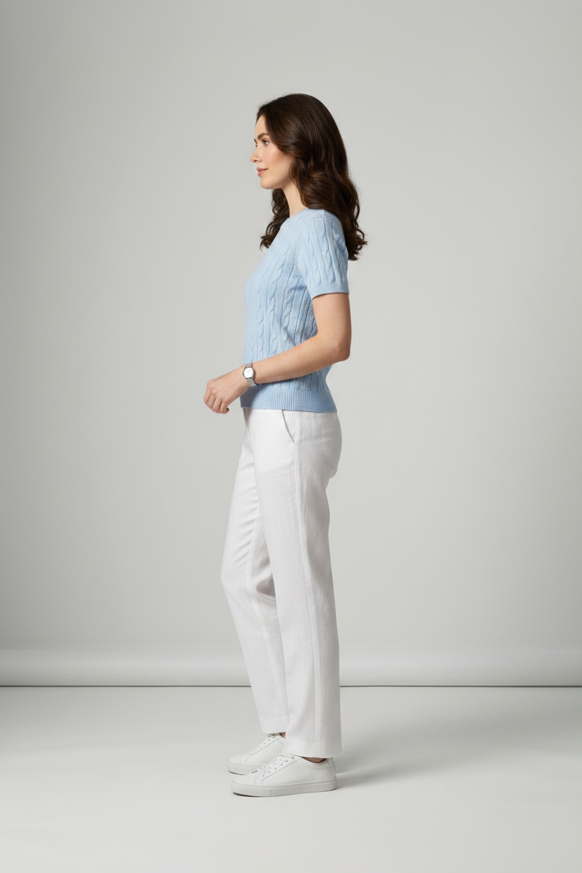 A woman stands in a studio wearing a light blue short-sleeve cable-knit sweater, white trousers, and white sneakers against a light gray background.