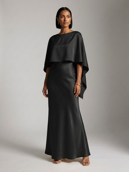 A woman modeling a floor-length black satin gown with a draped cape-style bodice and wide draped sleeves, photographed against a neutral light gray studio background.