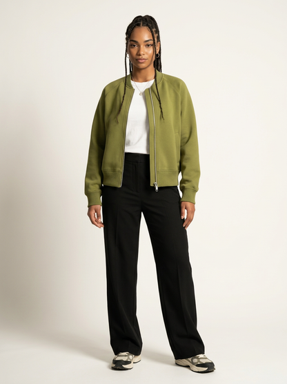 A model stands against a plain light beige studio background wearing an olive green zip-up bomber jacket over a white t-shirt, black wide-leg trousers, and gray sneakers.