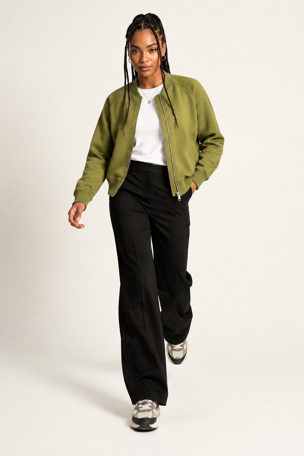 A woman modeling a cropped olive green bomber jacket over a white t-shirt and black wide-leg trousers, walking against a light gray studio background.