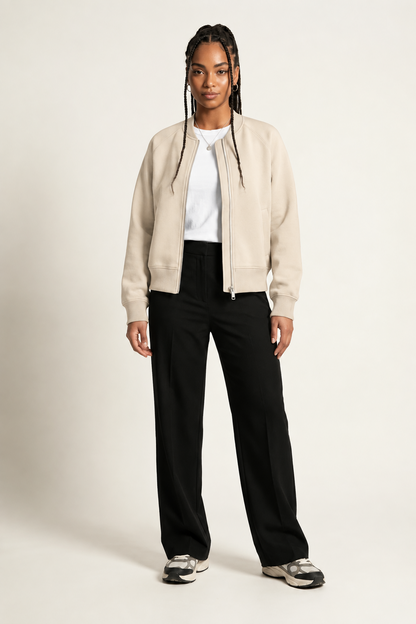 A model stands against a light beige studio background wearing a cream-colored zip-up bomber jacket over a white t-shirt, paired with black wide-leg trousers and gray sneakers.